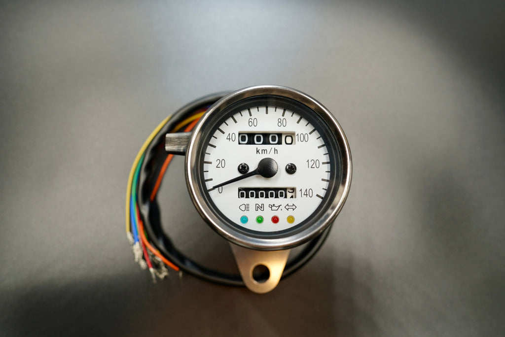 Retro analog motorcycle speedometer with white dial and stainless steel housing, shown front-on with indicator lights visible