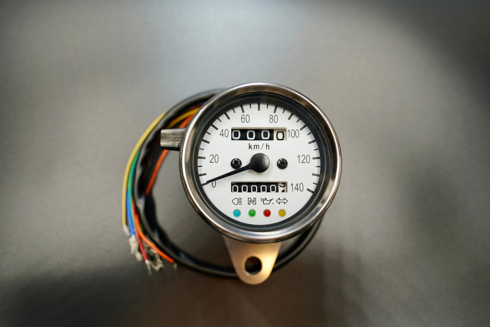 Retro analog motorcycle speedometer with white dial and stainless steel housing, shown front-on with indicator lights visible